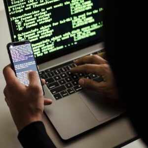 unrecognizable hacker with smartphone typing on laptop at desk